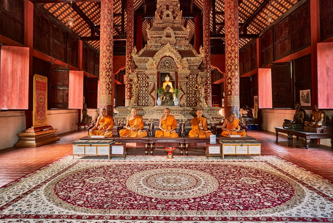 Buddhist monks in saffron robes seated in a traditional Thai temple with ornate golden altar and red wooden architecture.